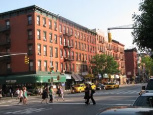 Pedestrians crossing a busy city street lined with historic red brick apartment buildings, storefronts, and yellow taxis.