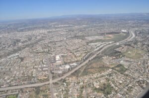 Aerial view of a densely populated suburban area in San Diego, California, featuring highways, residential neighborhoods, green spaces, and distant mountains under a clear blue sky.